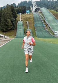 Sportoló szürke és fehér mintás sleeveless felsőt és rózsaszín részletekkel ellátott rövidnadrágot visel, és egy zöld műfűvön fut a síugró sáncok közelében.
