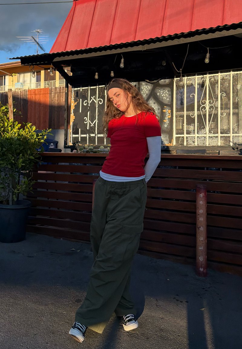 Jeune femme portant un t-shirt rouge, des manches longues blanches, un pantalon olive et un bandeau noir, posant en extérieur près d'une clôture en bois et d'une plante en pot sous la lumière du soleil.