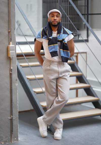 Camisa blanca de manga corta, pantalones beige ajustados y zapatillas blancas. Bufanda de rayas azules y negras colgada sobre los hombros. Entorno minimalista.