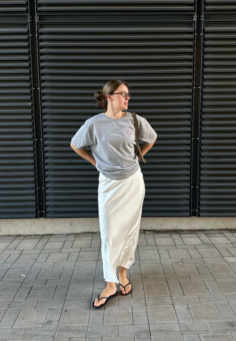 T-shirt cinza de algodão, saia maxi branca de cetim e chinelos pretos. Pose isolada contra um fundo texturizado com ripas escuras.