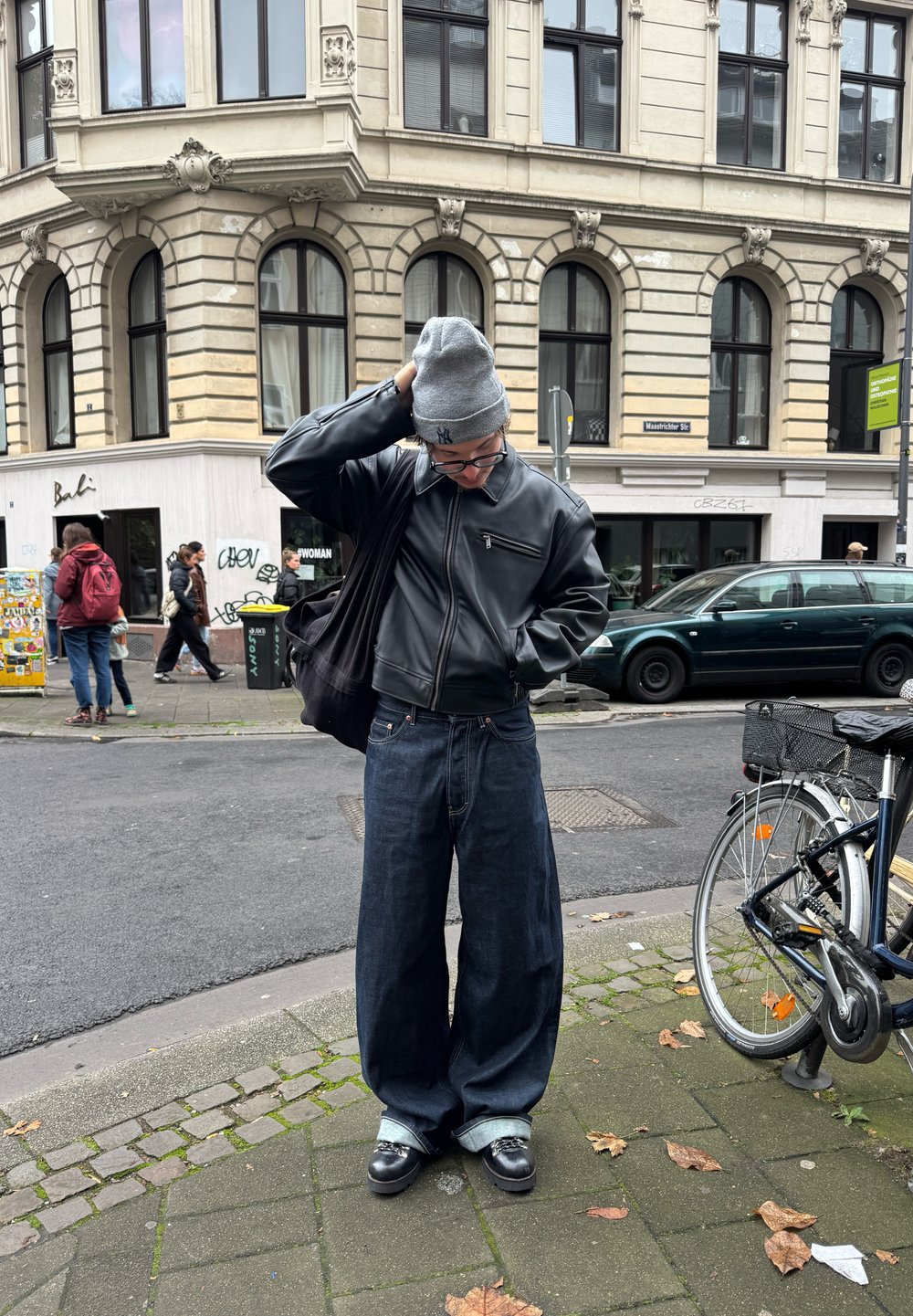Zwarte leren bomberjack, oversized blauwe denim jeans met omgeslagen pijpen, grijze gebreide beanie en zwarte schoenen. Stedelijke straatomgeving met fietsen.