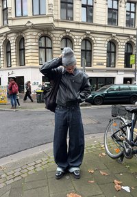 Zwarte leren bomberjack, oversized blauwe denim jeans met omgeslagen pijpen, grijze gebreide beanie en zwarte schoenen. Stedelijke straatomgeving met fietsen.