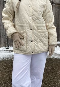 Giacca beige trapuntata con bottoni a pressione e tasche laterali, abbinata a guanti lavorati a maglia beige chiaro. I pantaloni bianchi completano l'outfit.