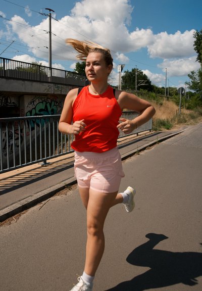Top atlético sin mangas rojo y pantalones cortos rosa claro, combinados con zapatillas blancas. Corriendo por un camino pavimentado con una pared graffiti de fondo.