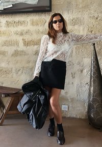 Čipkast zgornji del z dolgimi rokavi, črno teksturirana mini krilo, drži črno jakno. Temna očala, loaferji in nogavice. Nevtralna kamnita stena v ozadju.