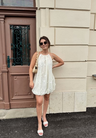 Vestido blanco sin mangas con bordado floral, dobladillo redondeado, combinado con sandalias trenzadas de color rosa claro. El fondo presenta una puerta marrón ornamentada.