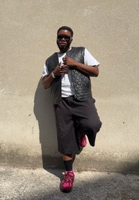 Černá prošívaná vesta přes bílé tričko, spárovaná s černými šortkami. Růžové a černé tenisky dotvářejí outfit. Osoba pózuje před zdí.