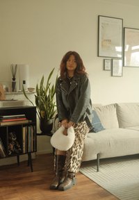 Svart lær bikerjakke, leopardmønstret skjørt, brune støvler til kneet. Holder en fluffy hvit håndveske. Innendørs miljø med planter og dekor.