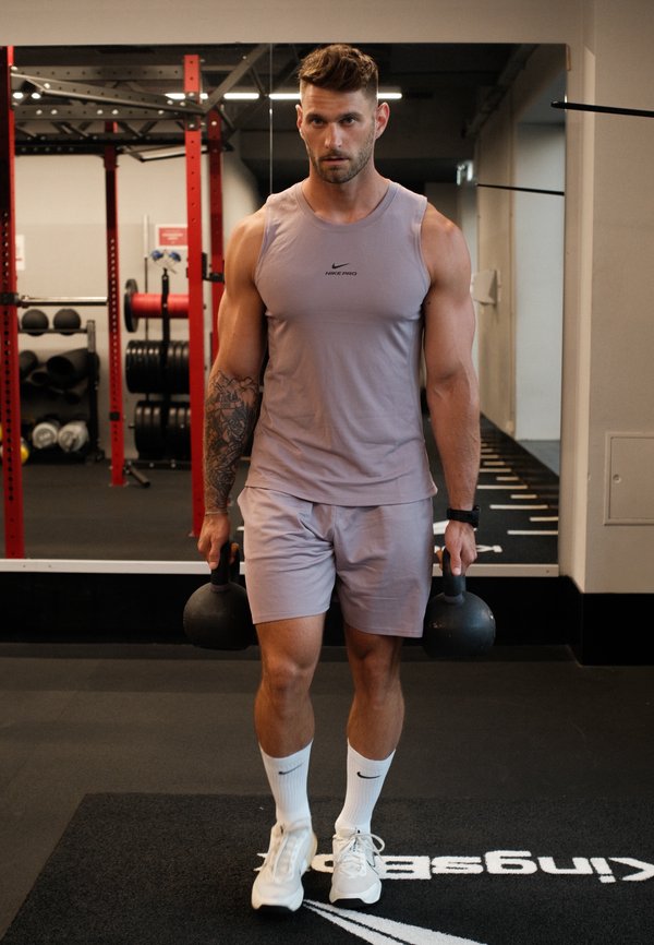 Homme musclé portant un débardeur et un short tient des kettlebells en marchant dans une salle de sport avec des poids et des racks en arrière-plan.