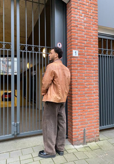 Chaqueta de cuero marrón, pantalones de pierna ancha marrón oscuro y zapatos negros, de pie cerca de una pared de ladrillos y una puerta de metal que conduce a un aparcamiento subterráneo.