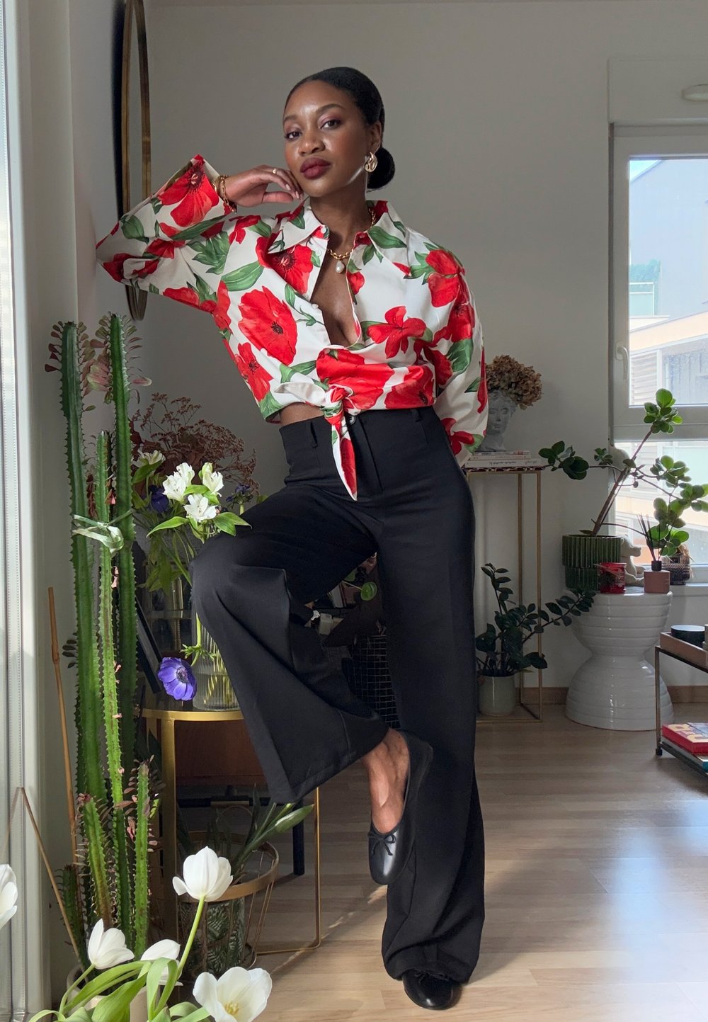 Camisa floral de botão com padrões vermelhos e verdes, amarrada na cintura, combinada com calças largas pretas e sapatos pretos. Fundo neutro.