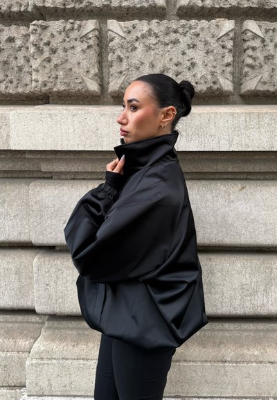 Mujer con cabello oscuro recogido en un moño ajusta el cuello alto de su chaqueta negra de gran tamaño, de pie de lado contra una pared de piedra texturizada.