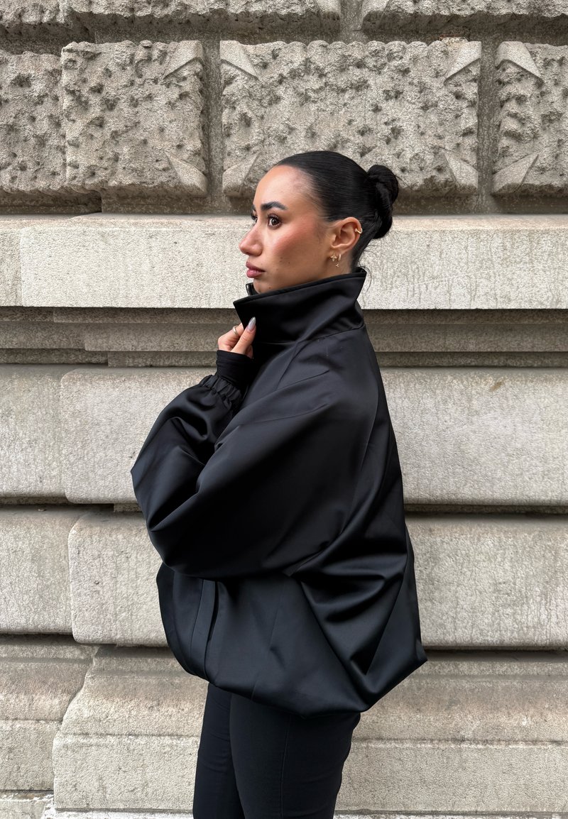 Femme aux cheveux foncés attachés en chignon ajuste le col haut de sa veste noire surdimensionnée, debout de profil contre un mur de pierre texturé.