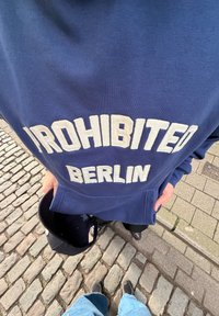 Personne portant un sweat-shirt bleu avec le texte « PROHIBITED BERLIN », tenant une casquette noire, debout sur un sol en pavés et en trottoir.