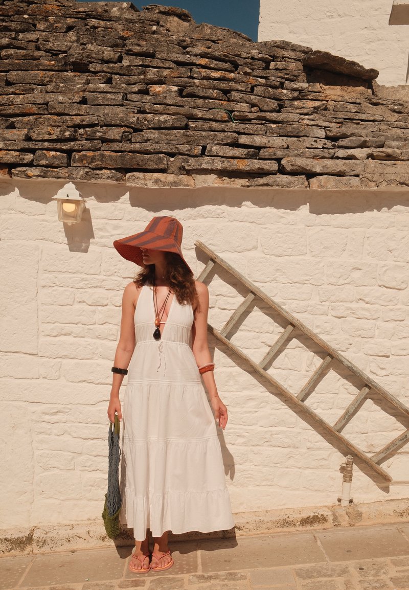 Femme en robe blanche et chapeau à large bord se tient contre un mur en pierre blanche avec une échelle en bois appuyée contre.