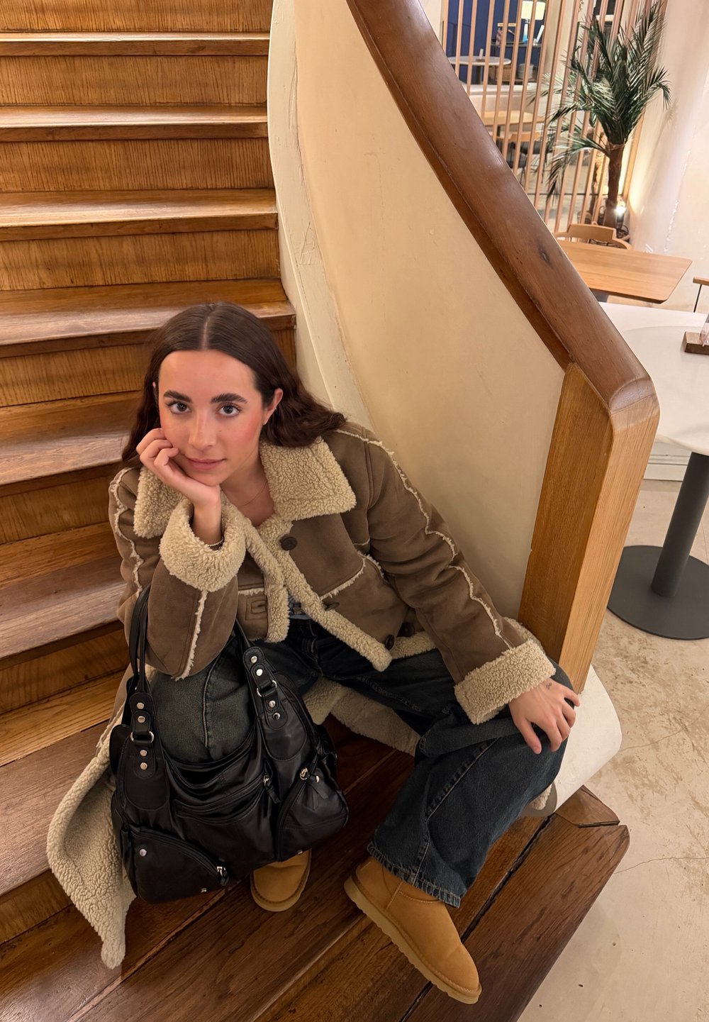 Jeune femme en veste en peau retournée et bottes assise sur des escaliers en bois à côté d'un sac noir dans un cadre de café chaleureux.