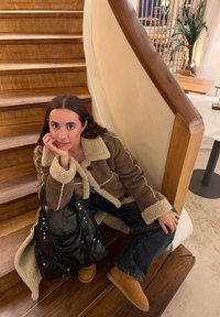 Jeune femme en veste en peau retournée et bottes assise sur des escaliers en bois à côté d'un sac noir dans un cadre de café chaleureux.