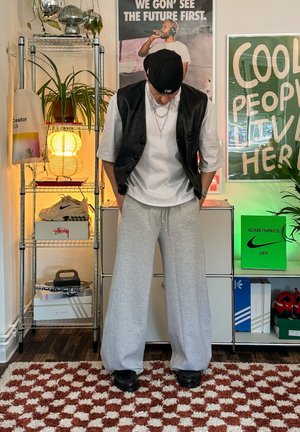 Chaleco de piel negro sobre una camiseta blanca, combinado con pantalones deportivos de pierna ancha grises y zapatos negros. El fondo presenta plantas y una decoración colorida.