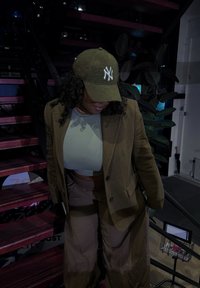 Hnědý blazer přes světlý top, spárovaný s oversized béžovými kalhotami; na hlavě má zelenou texturovanou kšiltovku s bílým logem, stojí na tmavých schodech.