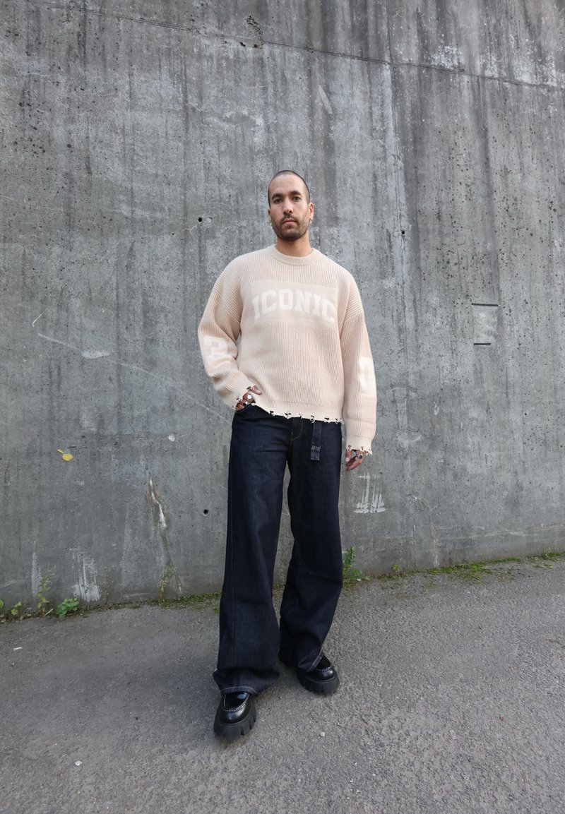 Pull en tricot beige surdimensionné avec le texte "ICONIC", associé à un pantalon large en denim foncé et des chaussures noires robustes. Détail de l'ourlet effiloché du pull.