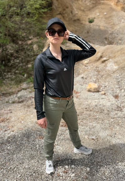 Camiseta atlética de manga larga negra con cuello con cremallera, pantalones cargo verdes, zapatillas grises y una gorra negra, en un fondo rocoso.