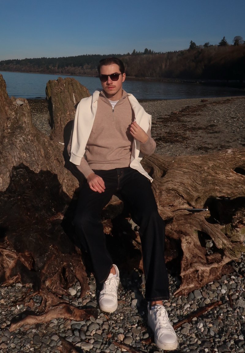 Hombre con gafas de sol, suéter beige y zapatillas blancas sentado sobre un gran trozo de madera a la deriva junto a una costa rocosa, con agua tranquila y un bosque al fondo.