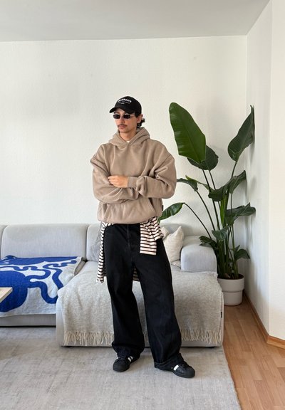 Hombre con sudadera con capucha beige, pantalones anchos negros, gorra y gafas de sol de pie en una sala de estar con sofá gris y planta alta en maceta.