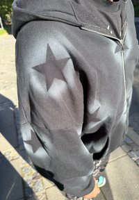 Persoană îmbrăcată cu un hanorac negru cu fermoar, împodobit cu modele de stele albe, stând în aer liber pe trotuarul pietruit luminate de soare.