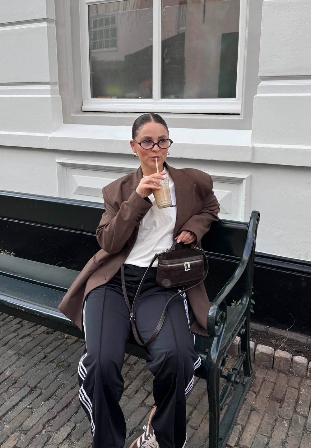 Brun oversized blazer, hvit t-skjorte, svarte treningsbukser med hvite striper, brun håndveske, runde briller, drikker iskald kaffe på en benk.