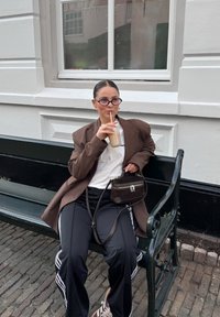 Brun oversized blazer, hvit t-skjorte, svarte treningsbukser med hvite striper, brun håndveske, runde briller, drikker iskald kaffe på en benk.