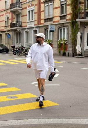 Camisa blanca de botones y pantalones cortos, zapatos negros, calcetines blancos, llevando una mochila negra. Calle urbana con paso de peatones amarillo y edificios de ladrillo.