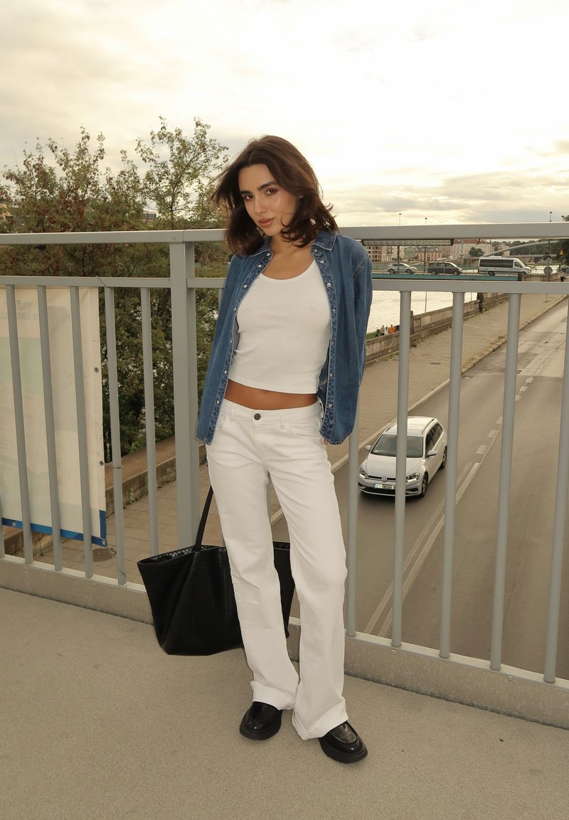 Camisa de ganga sobre um top curto branco, combinada com calças largas brancas e sapatos pretos chunky; segurando uma bolsa tote preta.