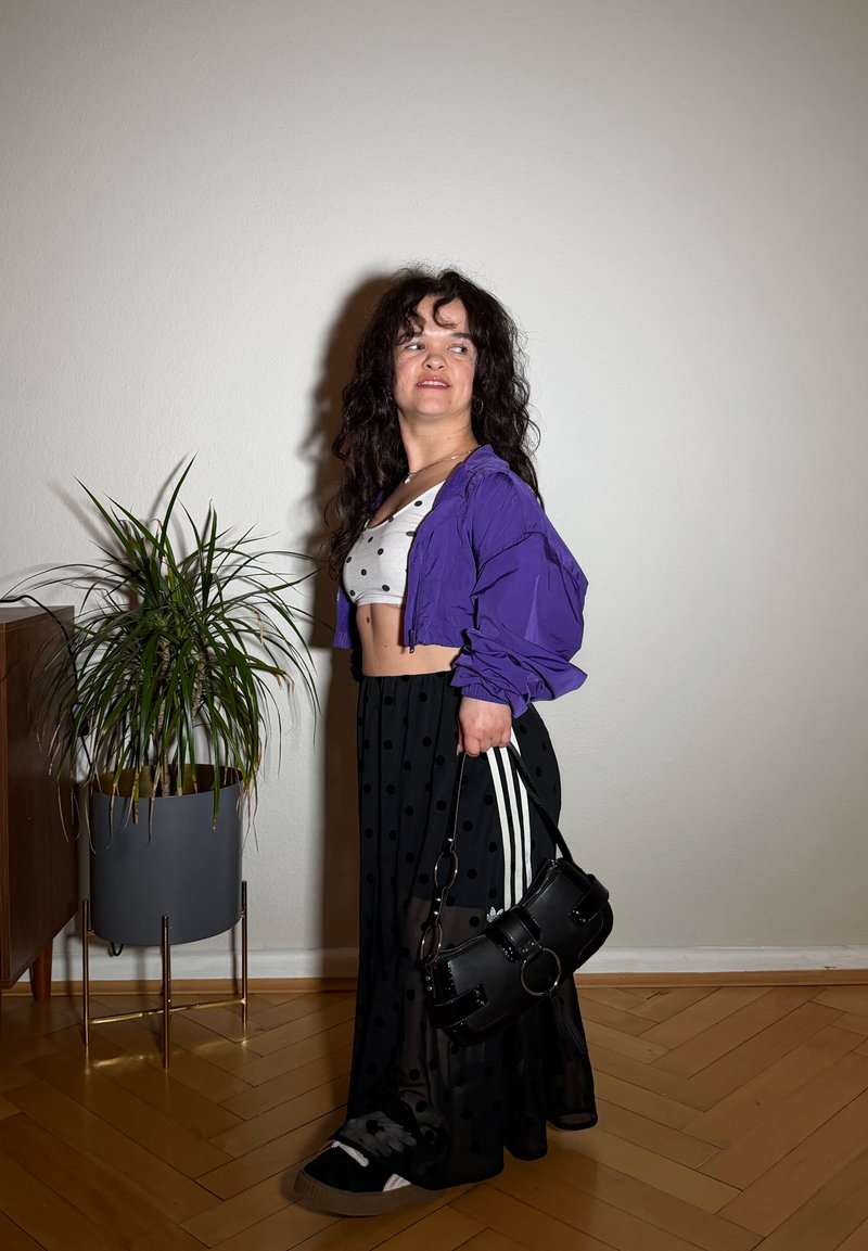 Femme aux cheveux bouclés portant une veste violette, un top court blanc et une jupe noire à pois, tenant un sac à main noir, debout près d'une plante en pot.