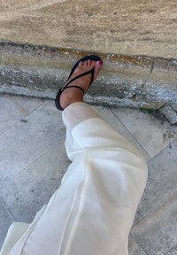 Schwarze Flip-Flop Sandale mit einem Riemen im Tanga-Stil, getragen zu hellen, lockeren Hosen, die einen lackierten Zehennagel und eine leichte Struktur des Stoffes zeigen.