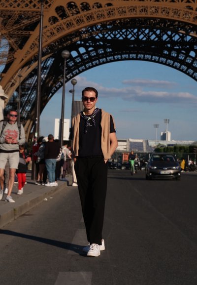 Chaleco beige sobre una camisa negra, pantalones negros y zapatillas blancas. El fondo presenta la estructura de rejilla de hierro de la Torre Eiffel.