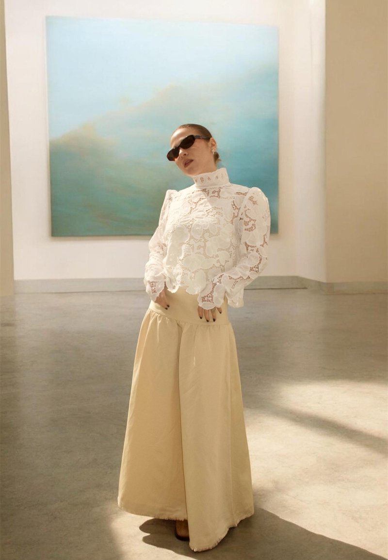 Woman wearing white lace blouse and wide beige pants poses indoors in front of a large blue and green abstract painting.