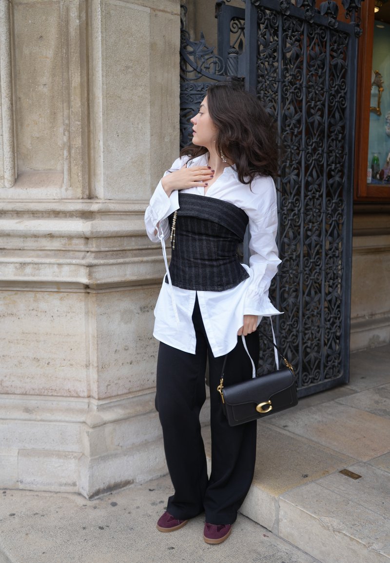 Corsetto nero, strutturato, sopra una camicia bianca; pantaloni neri a gamba larga; scarpe rosso scuro; borsetta nera piccola con dettagli in hardware dorato.