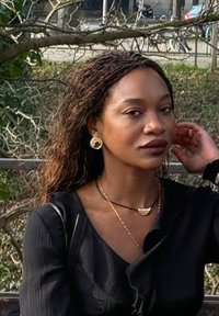 Femme aux cheveux foncés tressés, portant un haut noir et des bijoux dorés, assise en extérieur, touchant ses cheveux avec un parc et des branches d'arbres dénudées en arrière-plan.