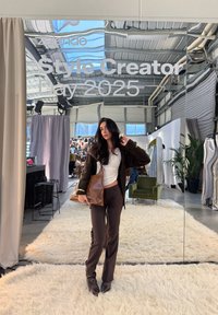 Brown oversized cardigan, white top, brown trousers, and leather handbag. Soft textured rug, mirrored background with event signage, indoor setting.