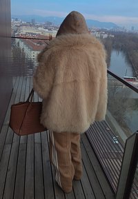 Personne en manteau de fourrure beige et pantalon rayé tenant un sac marron, debout sur un balcon surplombant une ville et une rivière avec des arbres dénudés.