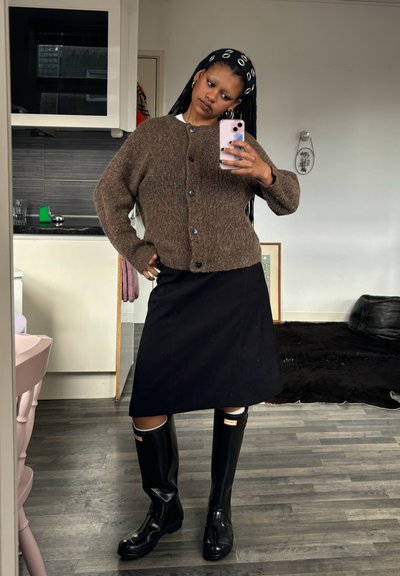 Cárdigan marrón de punto, falda negra hasta la rodilla y botas de goma negras brillantes. Cabello largo recogido con una cinta para la cabeza de estampado. Fondo neutro.