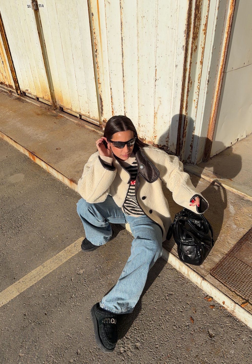 Jeune femme portant des lunettes de soleil, une veste crème, un pull rayé, un jean bleu clair et des chaussures noires, assise par terre près de portes métalliques rouillées, tenant un sac noir.