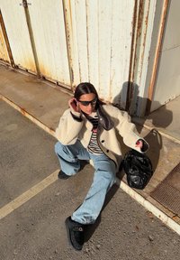 Jeune femme portant des lunettes de soleil, une veste crème, un pull rayé, un jean bleu clair et des chaussures noires, assise par terre près de portes métalliques rouillées, tenant un sac noir.