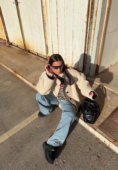 Joven mujer con gafas de sol, chaqueta color crema, suéter a rayas, jeans azul claro y zapatos negros, sentada en el suelo junto a unas puertas metálicas oxidadas, sosteniendo una bolsa negra.