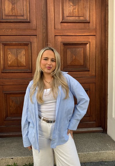 Joven con cabello largo y rubio que lleva una camisa azul clara sobre una camiseta blanca y pantalones blancos, de pie frente a una gran puerta panelada de madera.