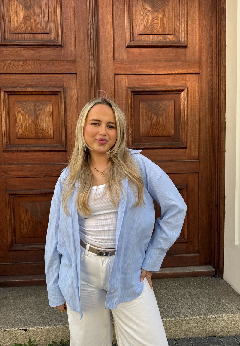 Jeune femme aux longs cheveux blonds portant une chemise bleu clair sur un top blanc et un pantalon blanc, se tenant devant une grande porte en bois à panneaux.