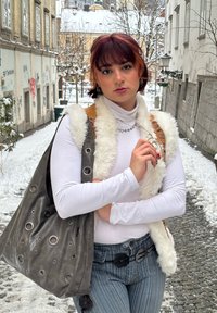 Sac à main en cuir gris avec des découpes circulaires, un col roulé blanc ajusté et un gilet en fausse fourrure. En arrière-plan, une rue enneigée et des bâtiments.