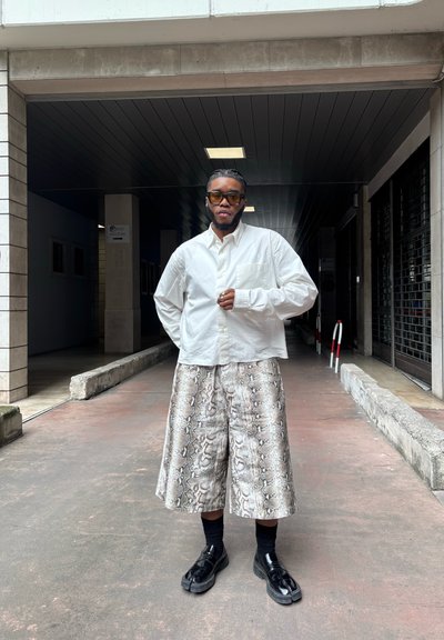 Camisa blanca de manga larga, pantalones cortos sueltos con patrón de serpiente plateado, calcetines negros y zapatos negros. El fondo presenta un pasillo cubierto.