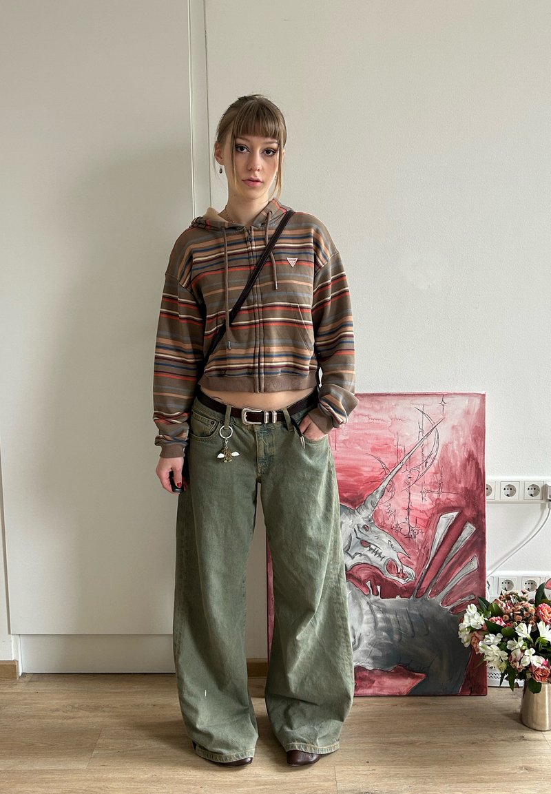 Persoană tânără stă în interior purtând un hanorac scurt cu dungi și pantaloni largi verzi, cu o pictură cu unicorn și un buchet de flori în fundal.