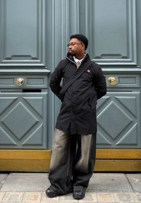 Black long jacket with a red and blue logo, oversized grey denim trousers, and black trainers. Standing in front of a teal door with gold hardware.